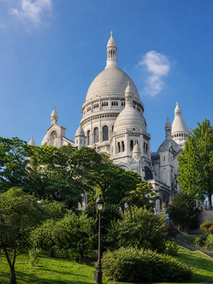 Die weißen Kuppeln der Sacré-Cœur-Basilika, die sich über die Bäume von Montmartre unter einem blauen Himmel erheben