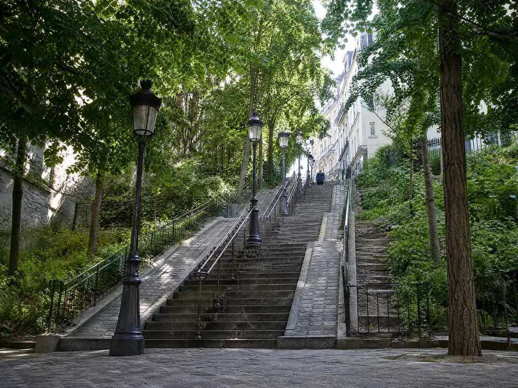 Lange Steintreppen mit Eisenhandläufen und Straßenlaternen, die einen grünen Montmartre-Hang unter Bäumen hinaufsteigen